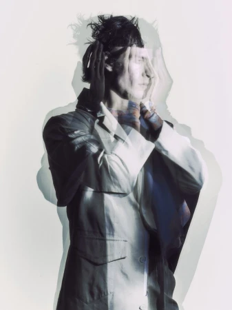 A man in a light tailored jacket posing with hands raised in a multi-exposure layered portrait against a pale studio backdrop.