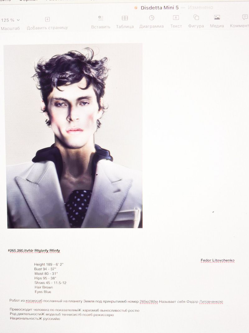 Portrait of a male fashion model in a white blazer with dark, wavy hair, posed in a photo studio setting with soft, diffused lighting.