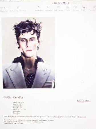 Portrait of a male fashion model in a white blazer with dark, wavy hair, posed in a photo studio setting with soft, diffused lighting.