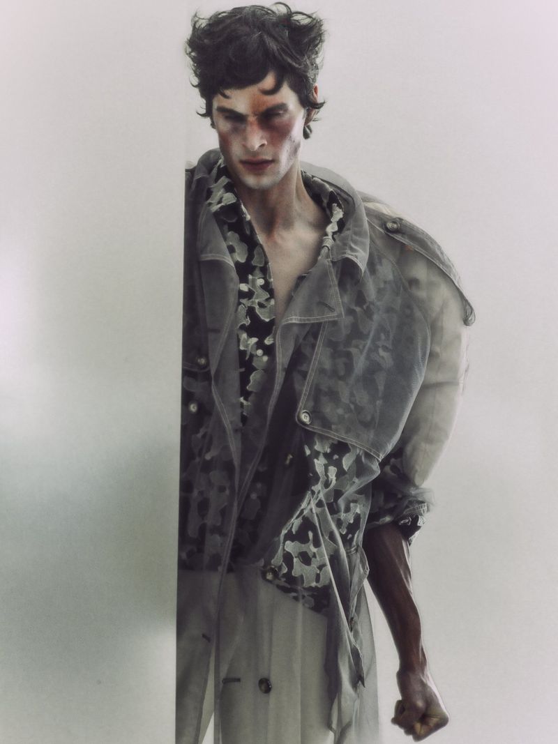 Portrait of a male model with an injured face in a gray jacket and patterned shirt against a plain white background, studio shot.
