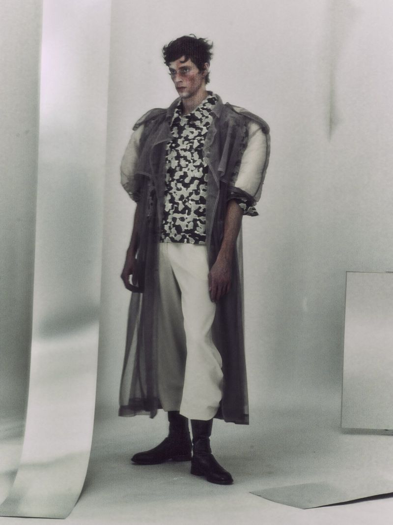 Fashionable adult man wearing a patterned shirt, long coat, wide leg trousers, and boots stands indoors in a studio setting.
