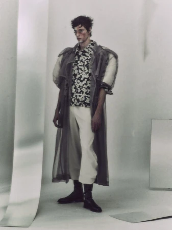 Man wearing a sheer coat, printed shirt, light trousers and dark boots standing in a minimalist studio setting.