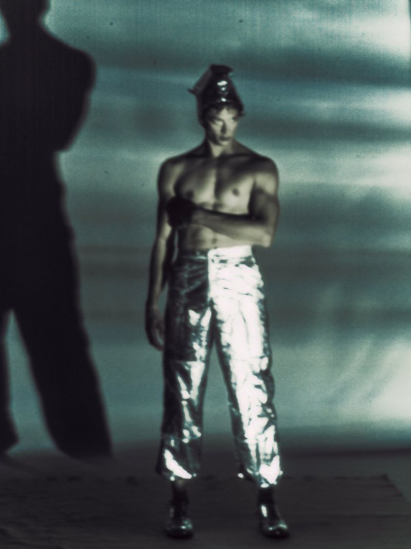Male fashion model poses in shiny pants and headwear in a studio against a blue and grey mottled background with his shadow visible.