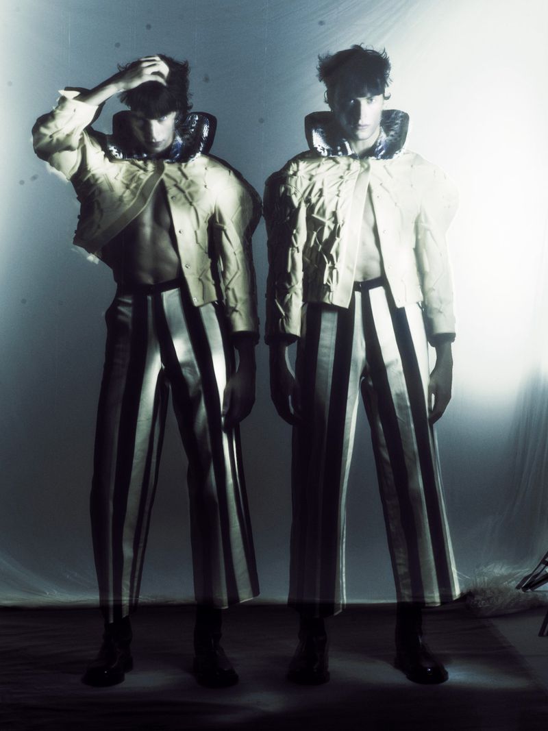 Two young adult males in striped pants and structured jackets pose in a studio with bright, diffused lighting for a fashion editorial.