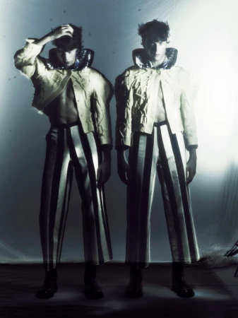 Two young adult males in striped pants and structured jackets pose in a studio with bright, diffused lighting for a fashion editorial.