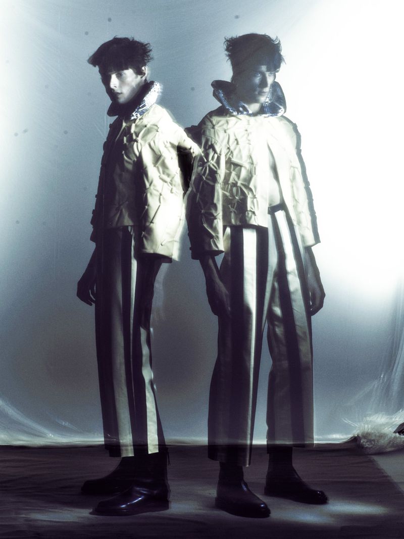 Two adult male models in an indoor studio, wearing matching avant-garde pants and jacket with white stripes, striking fashionable poses.