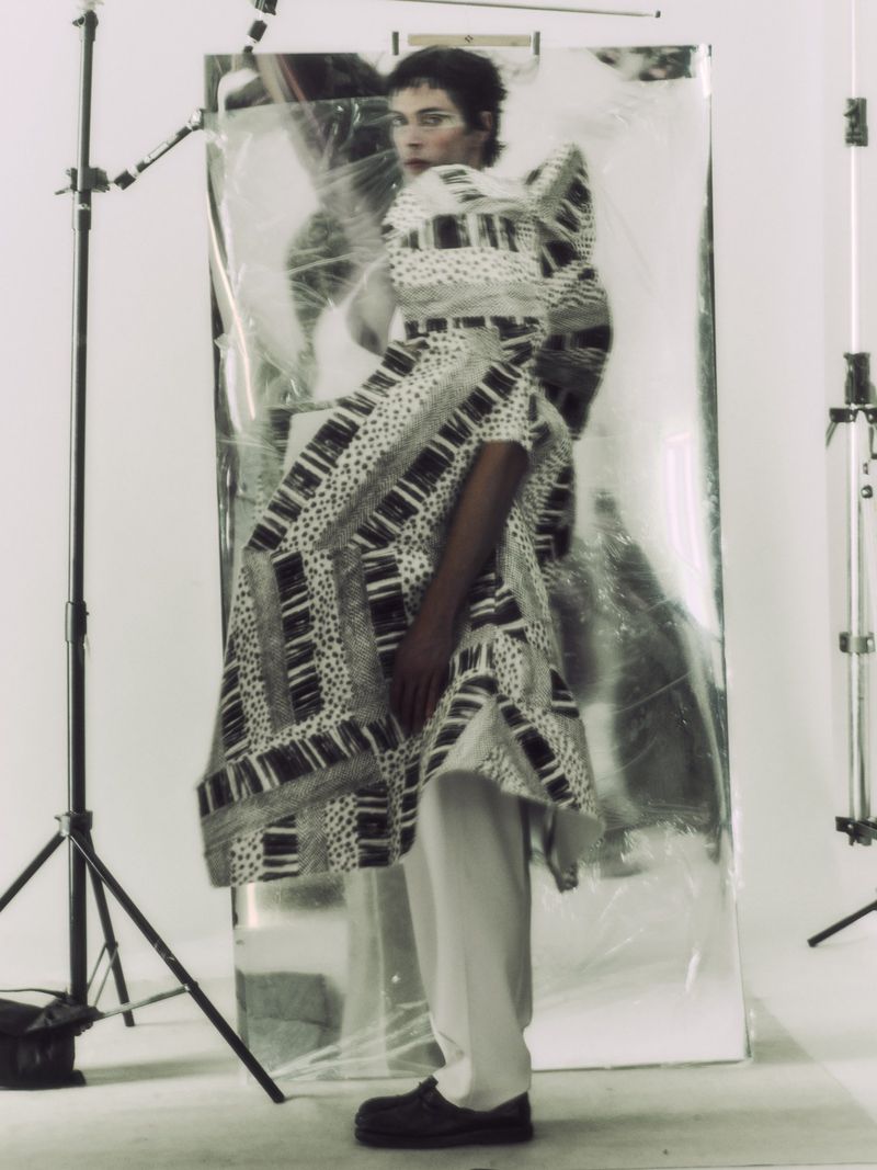 Model wearing a sculptural black and white patterned coat and pale trousers standing before a large metallic reflector in a studio.