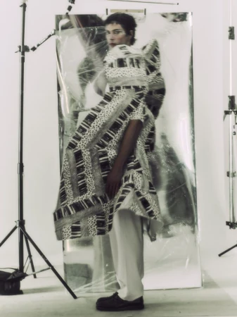 Model wearing a sculptural black and white patterned coat and pale trousers standing before a large metallic reflector in a studio.