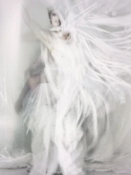 Motion blurred fashion portrait of a person draped in voluminous, ethereal flowing white fabric against a bright light background.
