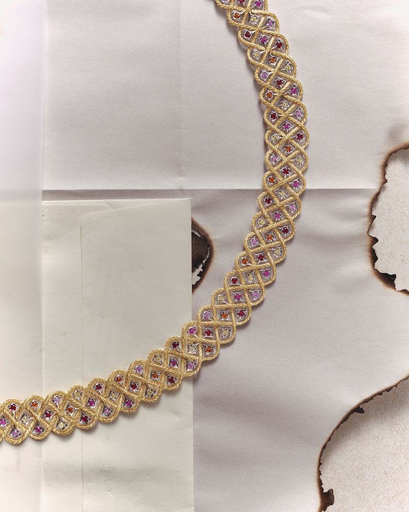 Braided gold necklace with pink and white stones, lying on a paper with burn marks, in a jewellery advertisement still life.