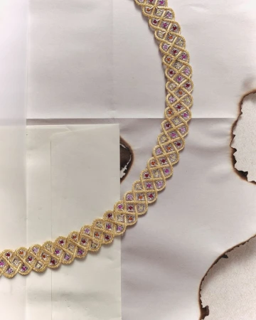 Braided gold necklace with pink and white stones, lying on a paper with burn marks, in a jewellery advertisement still life.
