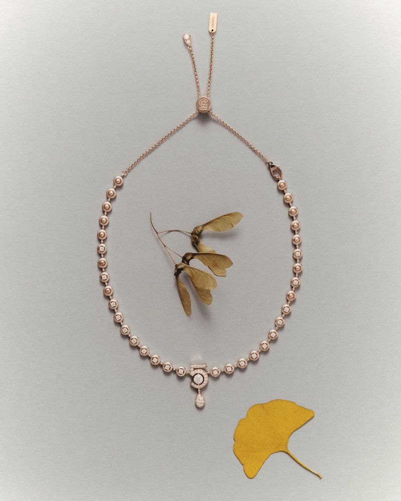 Elegant rose-gold necklace with pendant and crystal accents, styled with maple leaves on a light-grey surface. Jewellery product shot.