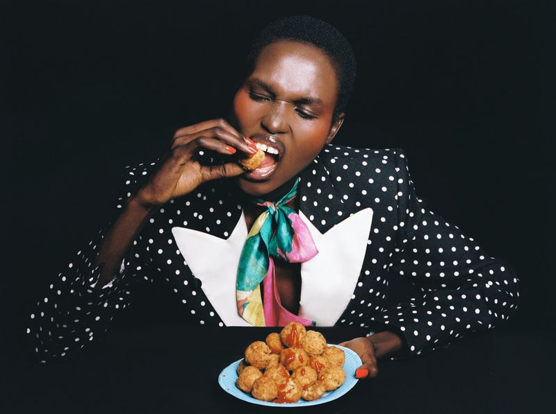 Woman with short dark hair in polka dot blazer eats fried food covered with red sauce from a blue plate in a studio setting.