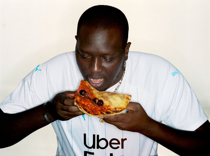A person with dark skin eats a slice of pizza. They are wearing a white shirt and silver chain, set against a solid white background.