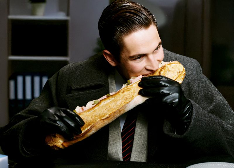 A man in a suit and gloves is eating a large sandwich. He is indoors with gray walls and dark filing cabinets in the background.