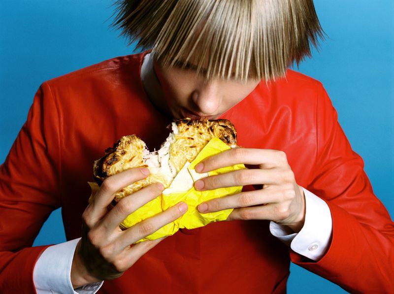 Person in red jacket eating a large sandwich with white filling wrapped in yellow paper against a blue background in a studio.