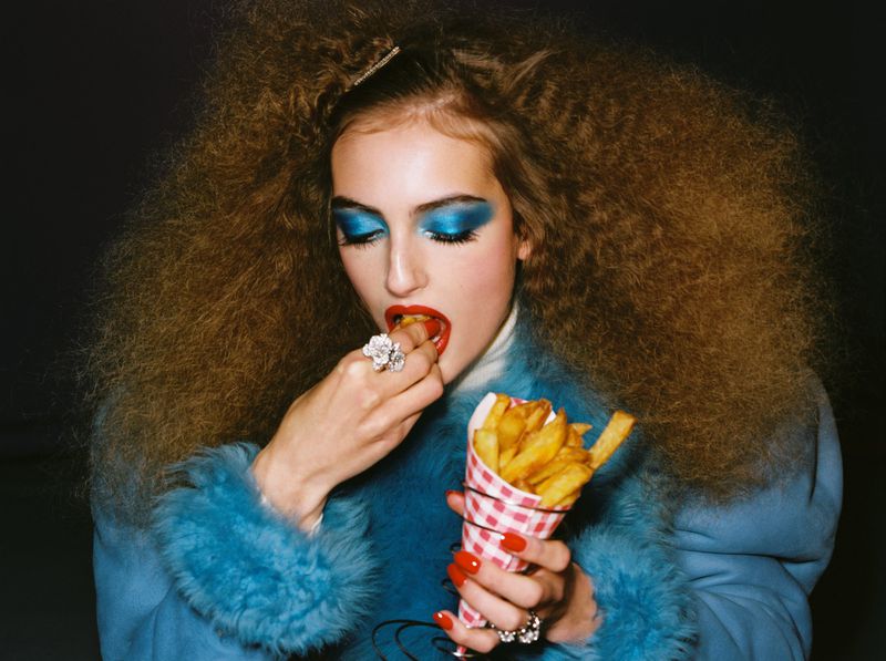 Woman with curly hair and bold makeup is eating french fries from a checkered paper cone while wearing a fuzzy, turquoise jacket.