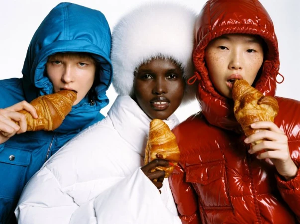Three diverse models in puffy coats eat croissants against a white backdrop, showcasing bright fashion and simple food indulgence.