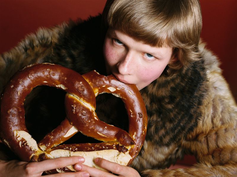 A person in a furry coat is biting into a large, fresh pretzel. The studio shot features a warm red backdrop and simple composition.