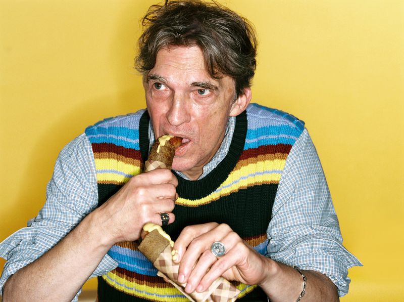 Man with short brown hair and a striped sweater vest is eating a wrap with a yellow filling, posing against a yellow backdrop.