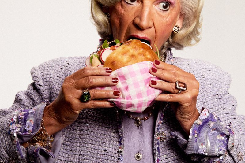 Elegant senior woman in a lavender suit jacket eating a pita sandwich with a shocked expression on a white background in a studio.