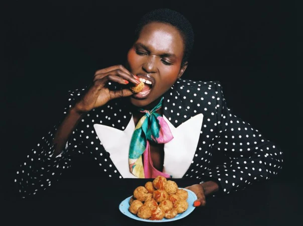 Model Amar Akway wearing a polka dot blazer and silk neck scarf eating fried snacks from a blue plate against a black background.
