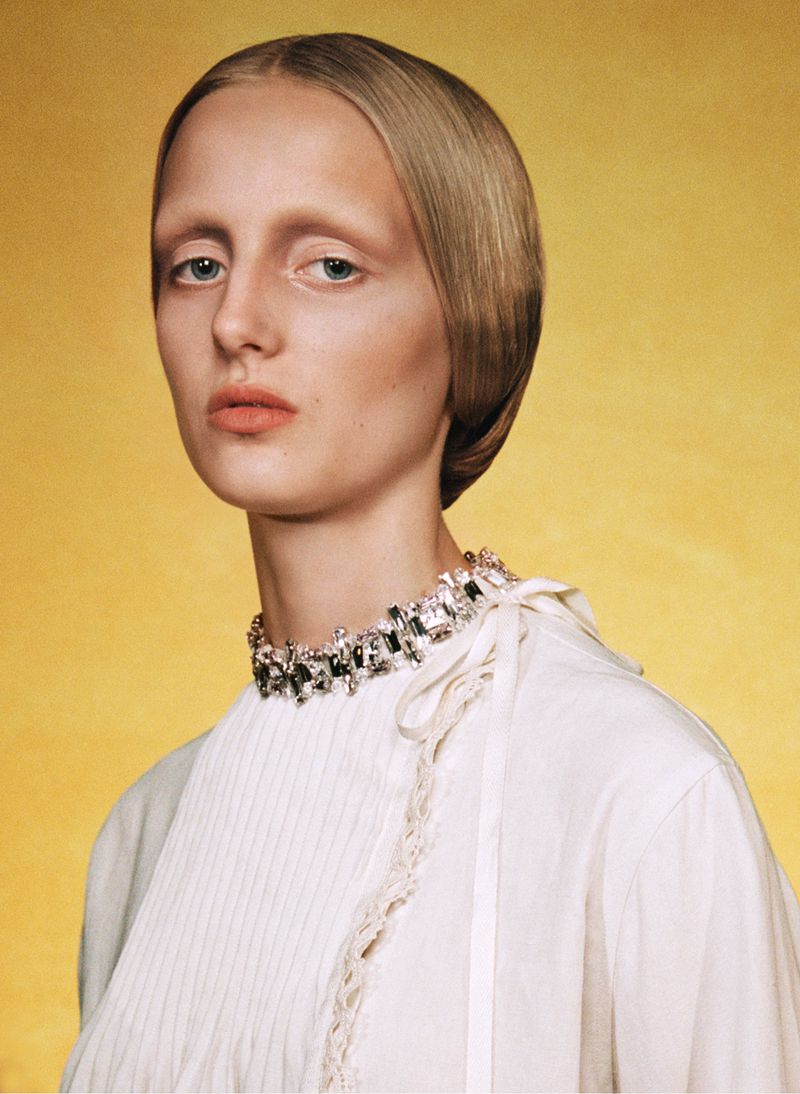 Woman with slicked-back blonde hair and neutral expression posing against a yellow backdrop wearing a jeweled choker and white top.