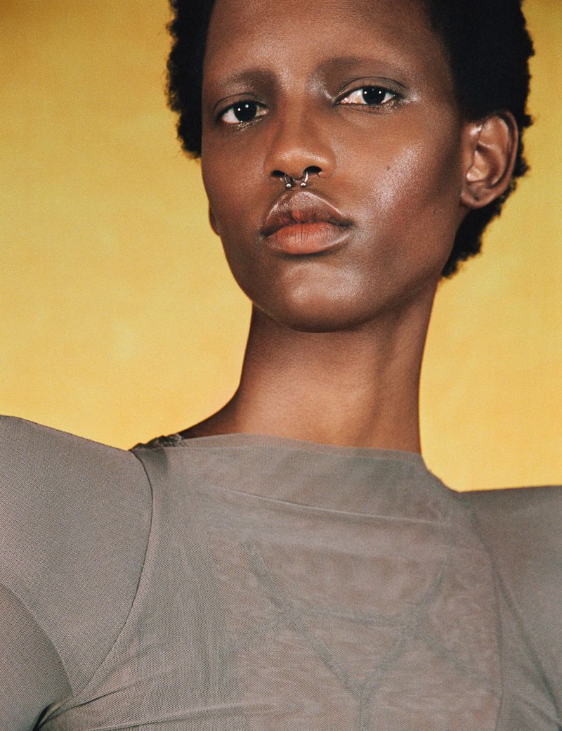 Portrait of an African American woman with dark hair and septum piercing, wearing a grey top against a mottled yellow background.