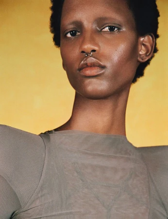 Portrait of an African American woman with dark hair and septum piercing, wearing a grey top against a mottled yellow background.