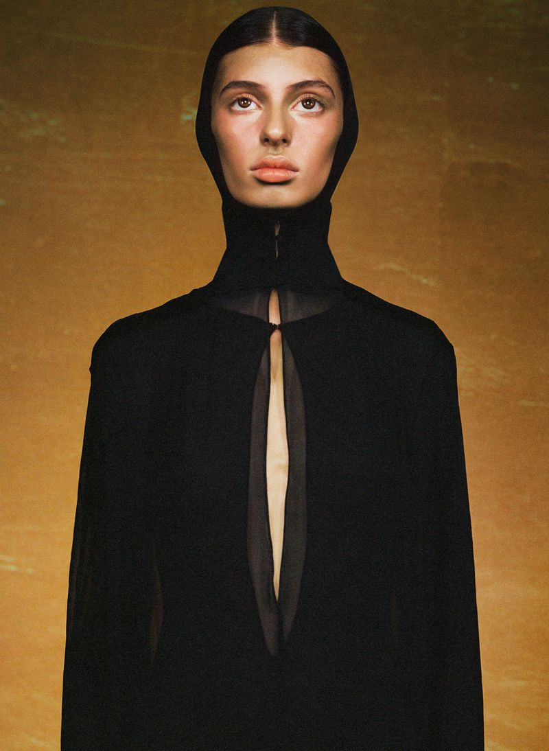 Close-up studio shot of a woman with a neutral expression, wearing a dark hooded garment against a textured gold background.
