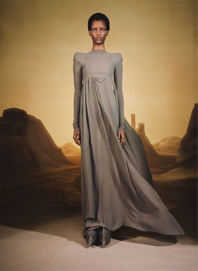 An African American woman showcases a long-sleeved grey gown against a surreal background of soft dunes and distant castle.