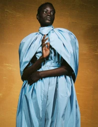 Model posing in a voluminous powder blue caped dress against a shimmering golden textured backdrop.