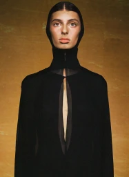 Model in a black hooded high-neck garment with a center keyhole slit against a warm textured gold-toned studio background.