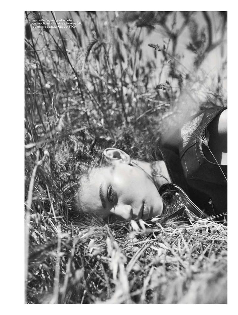 Monochrome shot of a woman lying in tall grass, eyes closed and wearing a dark vest. The background is filled with out-of-focus grass.