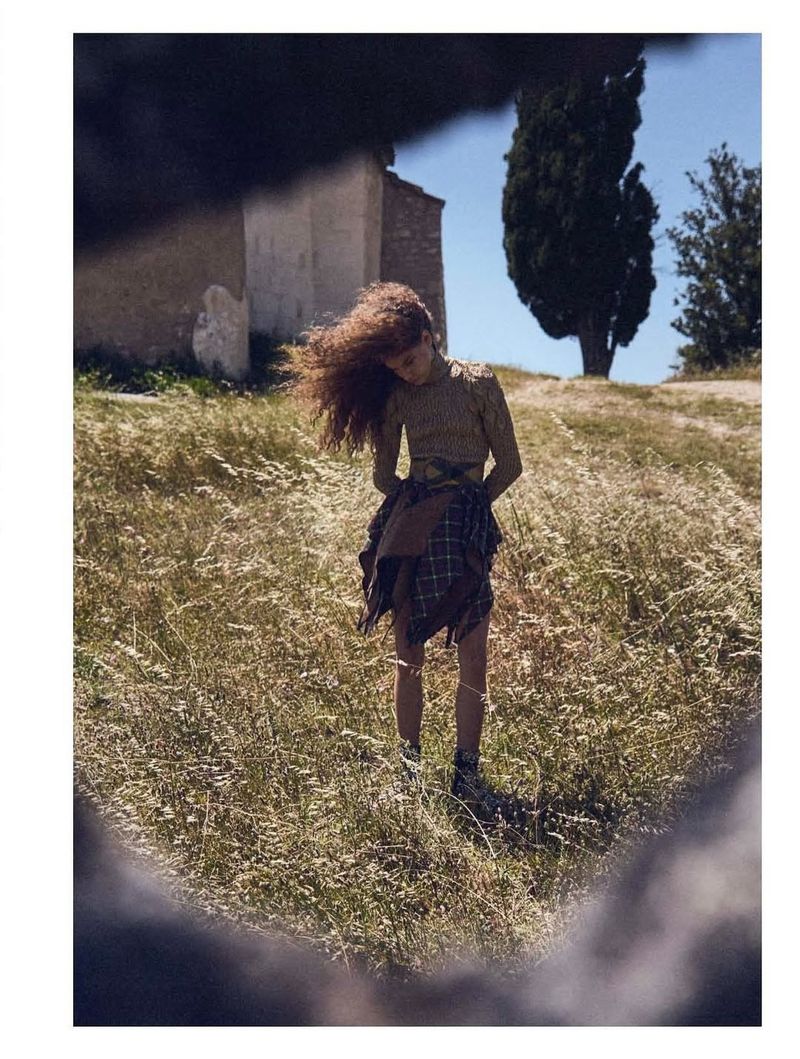 Red-haired woman stands in a grassy field with a stone building and tree in the background on a sunny day, outdoor fashion editorial.