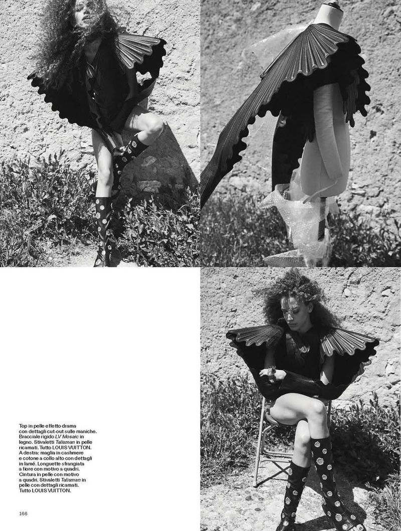 Black and white fashion editorial shot outdoors. A model poses in avant-garde clothing near a wall and grass in natural light.
