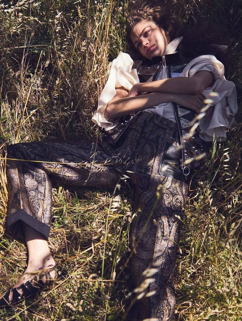 Woman lies in grassy field in an ornate pants and a peasant-style blouse, captured in natural outdoor lighting with eye-level framing.