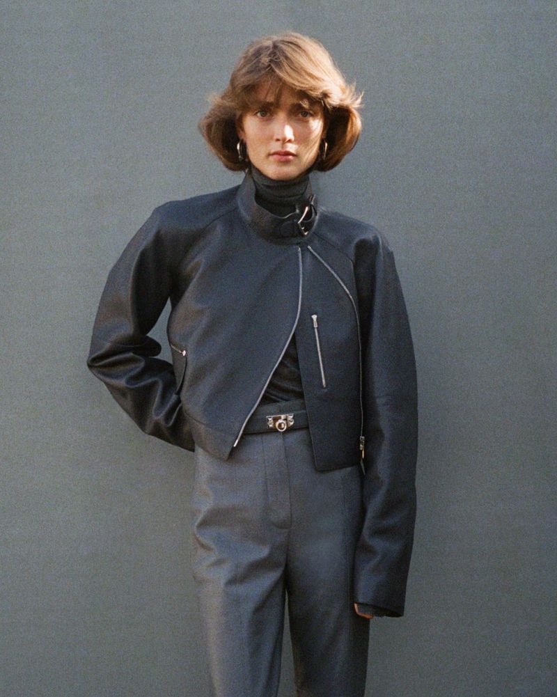 Woman in a black leather jacket and gray pants, posing in front of a plain gray wall in natural sunlight, fashion portrait.