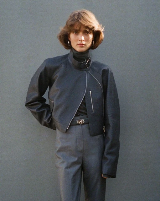 Woman in a black leather jacket and gray pants, posing in front of a plain gray wall in natural sunlight, fashion portrait.