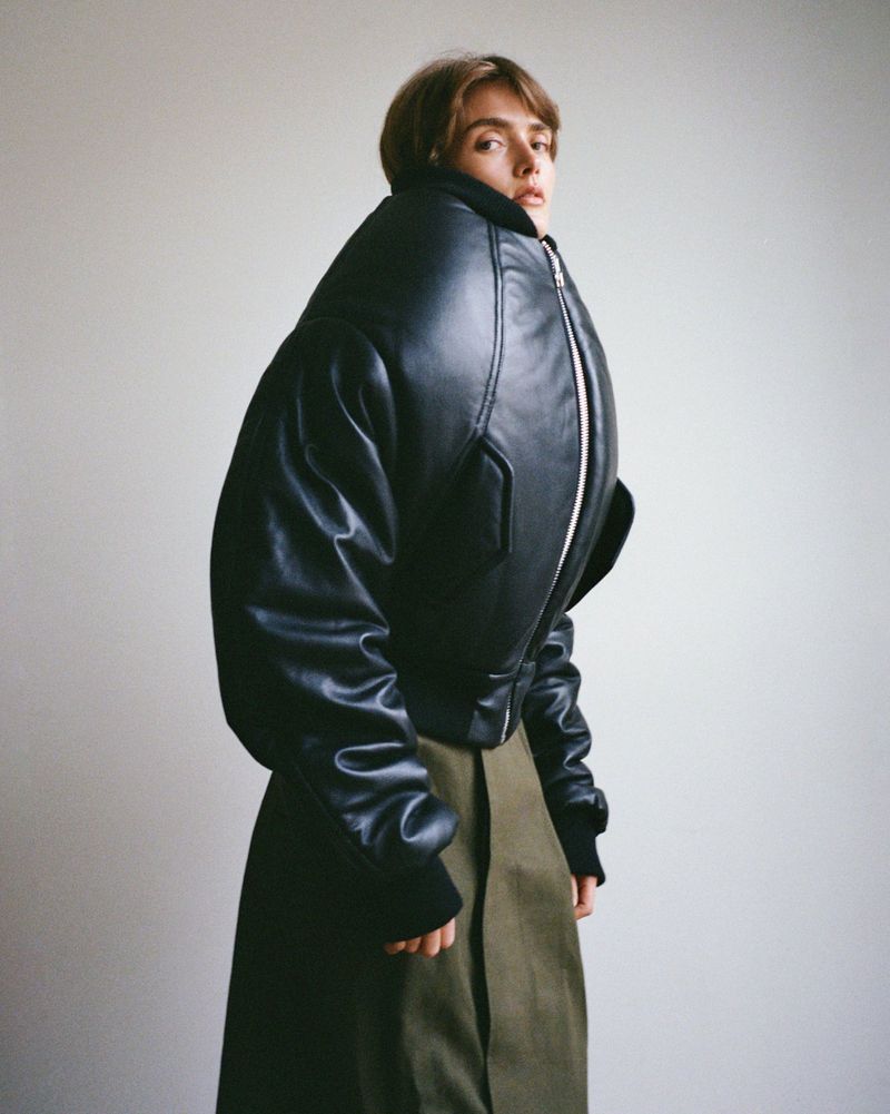A single adult woman in a black, oversized bomber jacket and green skirt stands against a plain white studio backdrop in a fashion shot.
