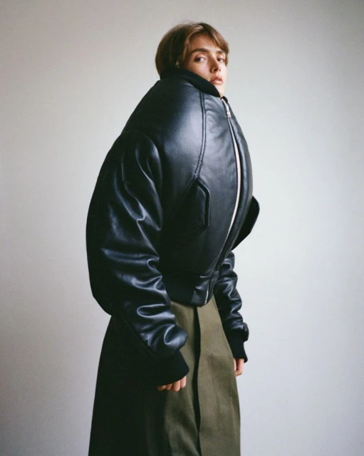A single adult woman in a black, oversized bomber jacket and green skirt stands against a plain white studio backdrop in a fashion shot.