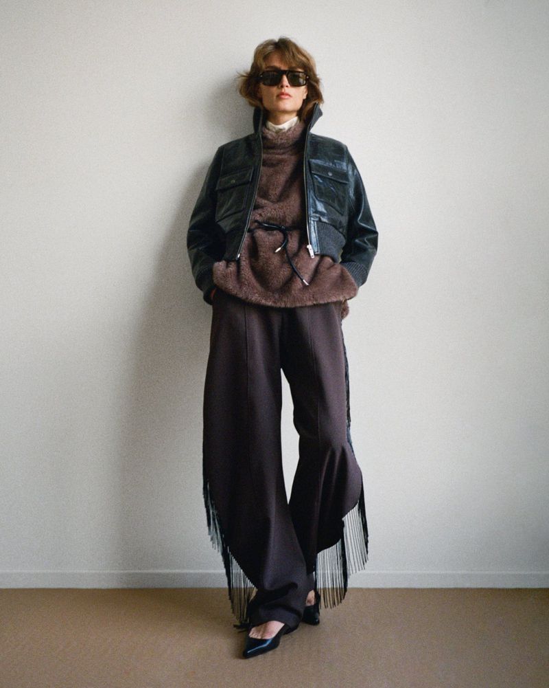Fashionable woman poses in a black leather jacket, brown top, and wide-leg brown pants with fringe against a neutral wall.