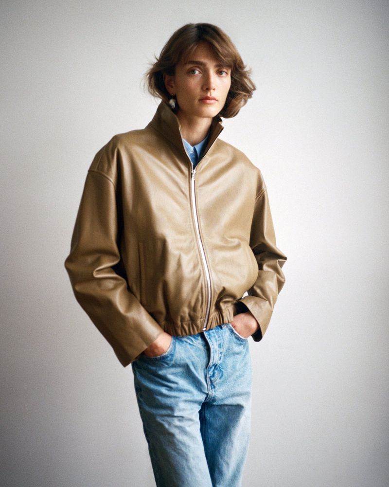 Woman in a studio setting wearing a tan leather jacket and light blue jeans. The image is a medium shot with a white background.
