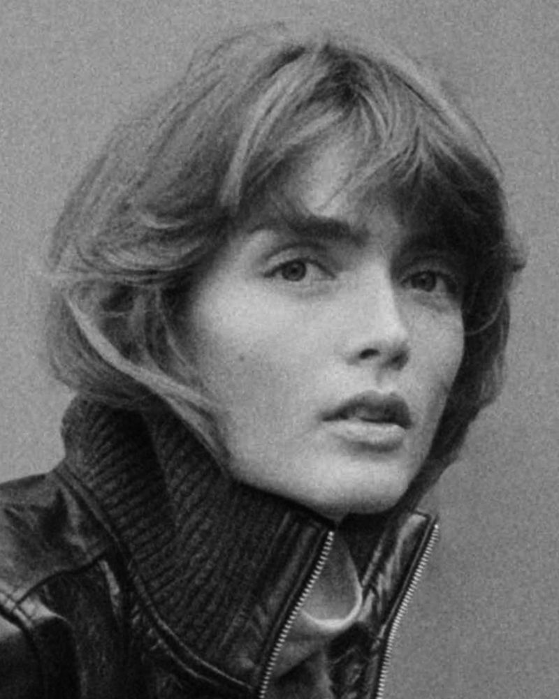 Black and white close-up portrait of a woman with feathered hair wearing a high-collared leather jacket against a grey background.
