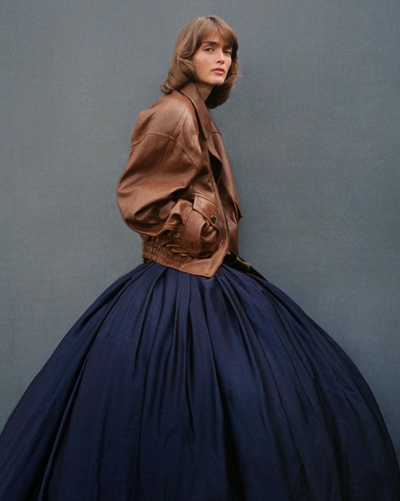 Fashionable woman poses wearing a brown leather bomber jacket and large navy blue skirt against a grey wall in a studio setting.