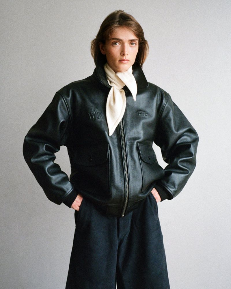 Fashion portrait of a female model in a black leather bomber jacket and cream scarf against a neutral studio backdrop. Modern, chic.
