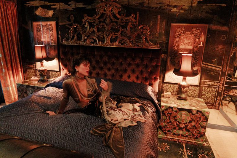 Woman lounges on an ornate bed with a velvet headboard in an opulent bedroom with side tables and lamps under warm, moody lighting.