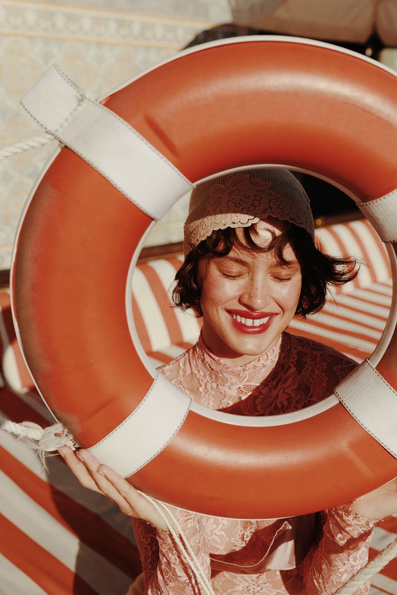 Woman in lace dress and hat framed by orange life preserver with red and white striped background in a close-up eye-level outdoor shot.