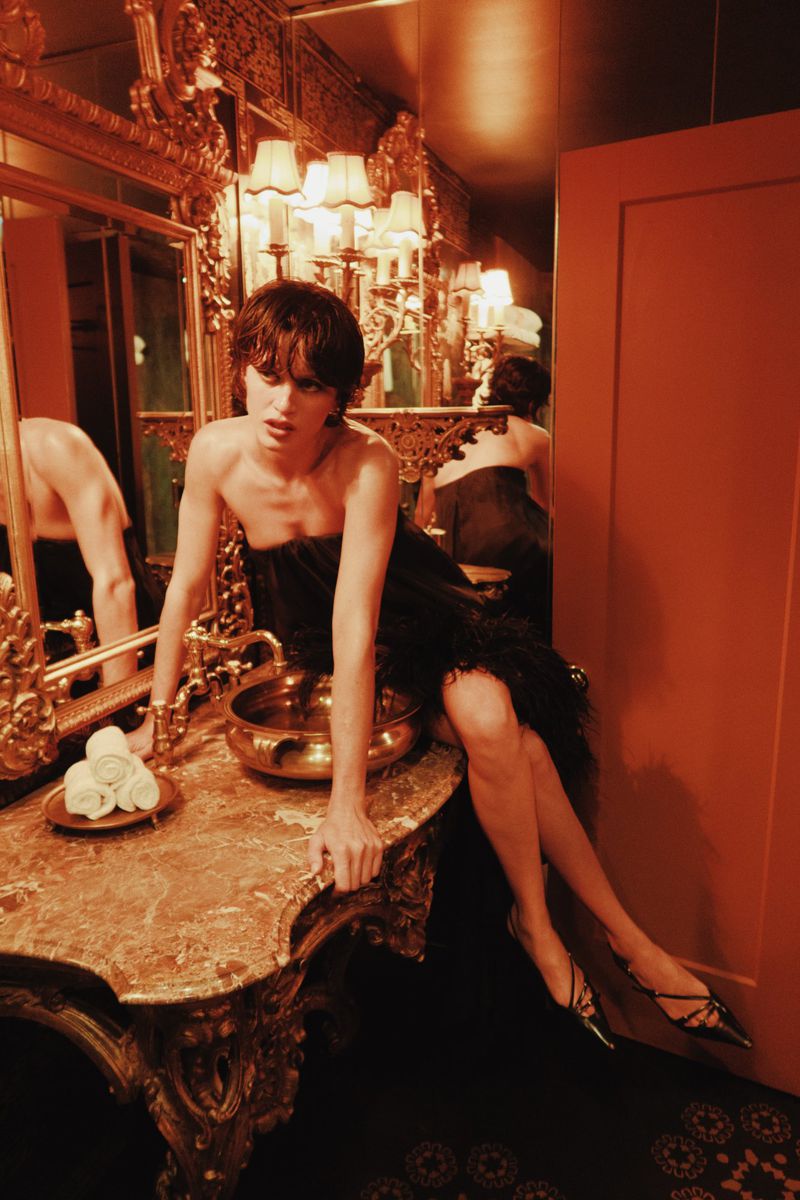 Woman in a black dress poses in a bathroom with an ornate gold vanity, faucet and mirror. The lights are warm and ambient.