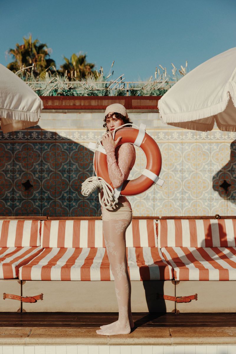 Woman in swimwear with a lifesaver ring stands near an outdoor swimming pool with striped seating and patterned tile background.